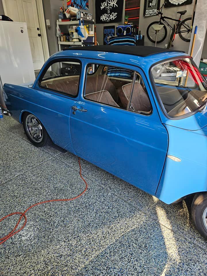 A bright blue vintage car parked inside a garage with tan interior seats and a black roof panel.