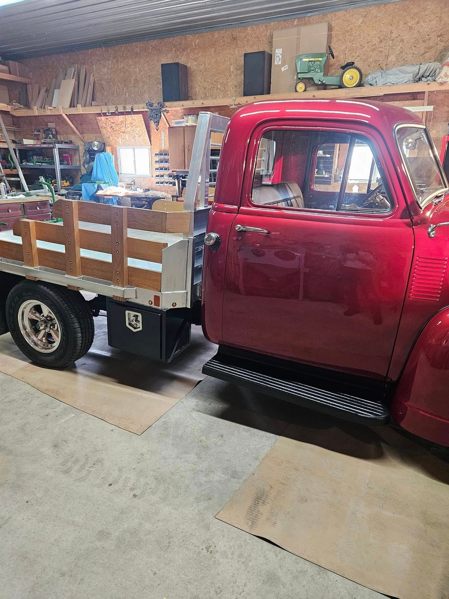 A bright red vintage pickup truck with a wooden flatbed parked inside a workshop with tools and shelving in the background.