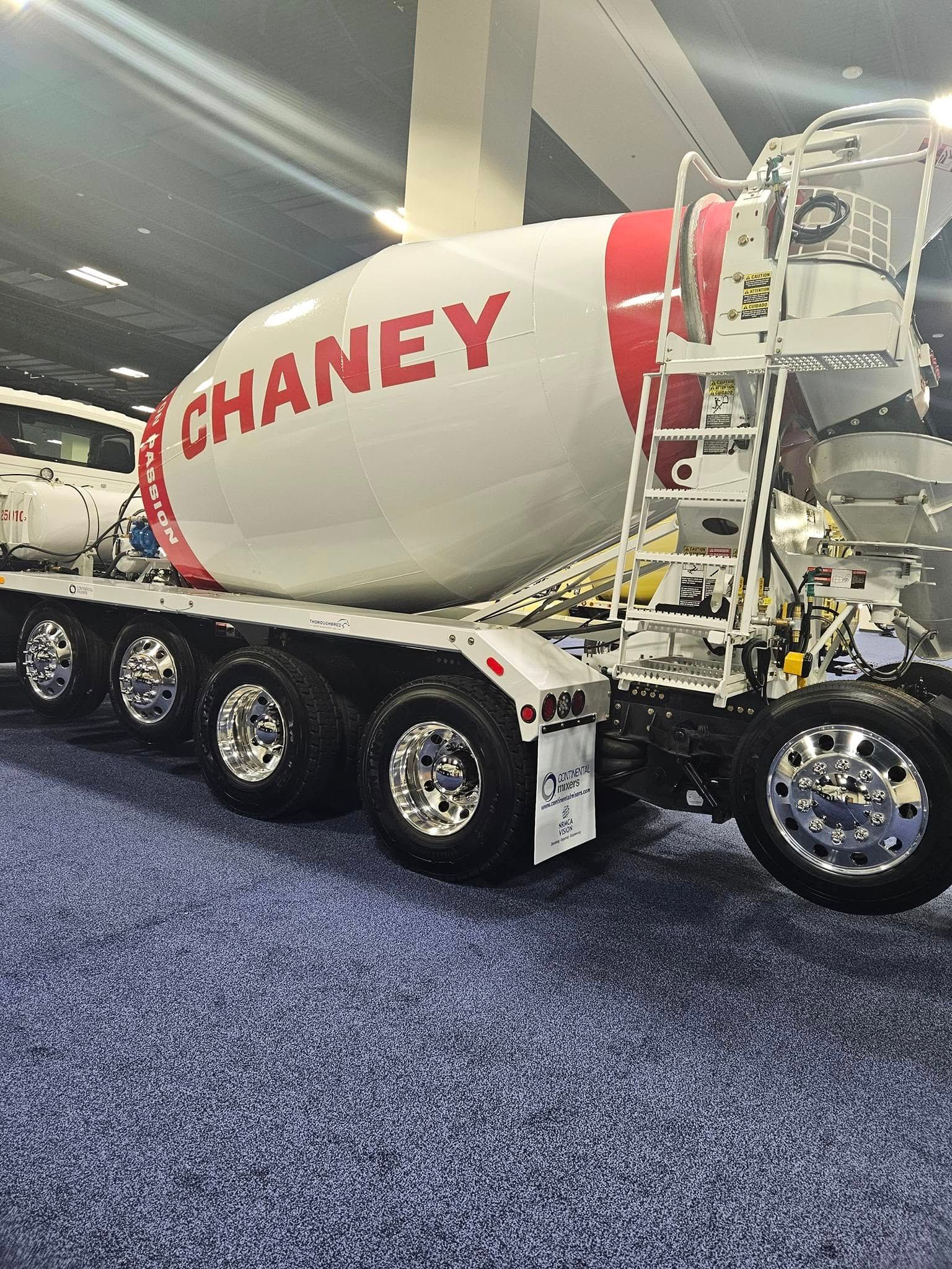 A white and red Chaney concrete mixer truck displayed on a blue carpeted floor at an indoor event.