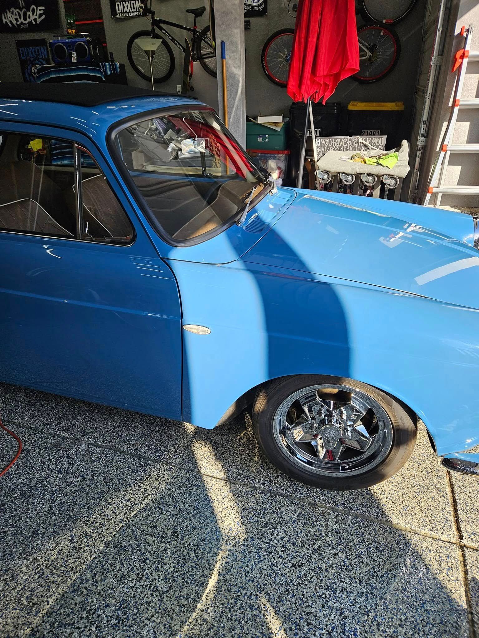 A bright blue, lowered vintage car parked inside a garage with bicycles and equipment visible in the background.