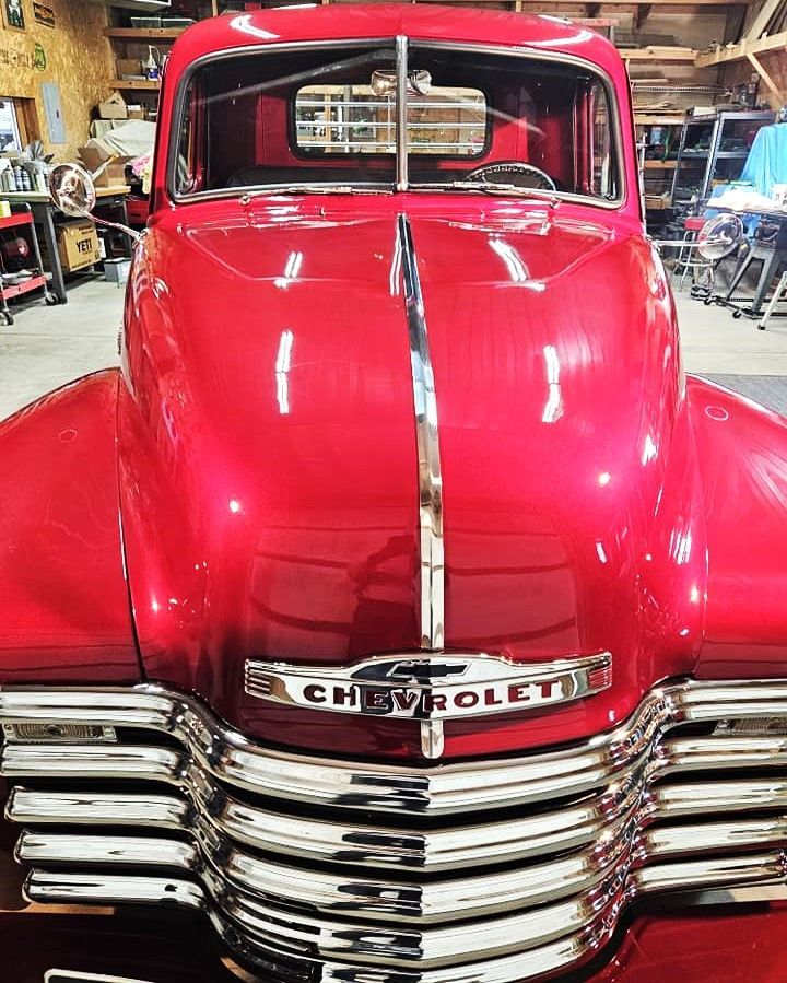 A bright red, vintage Chevrolet truck, front-facing, with a prominent chrome grille, parked in a cluttered garage.