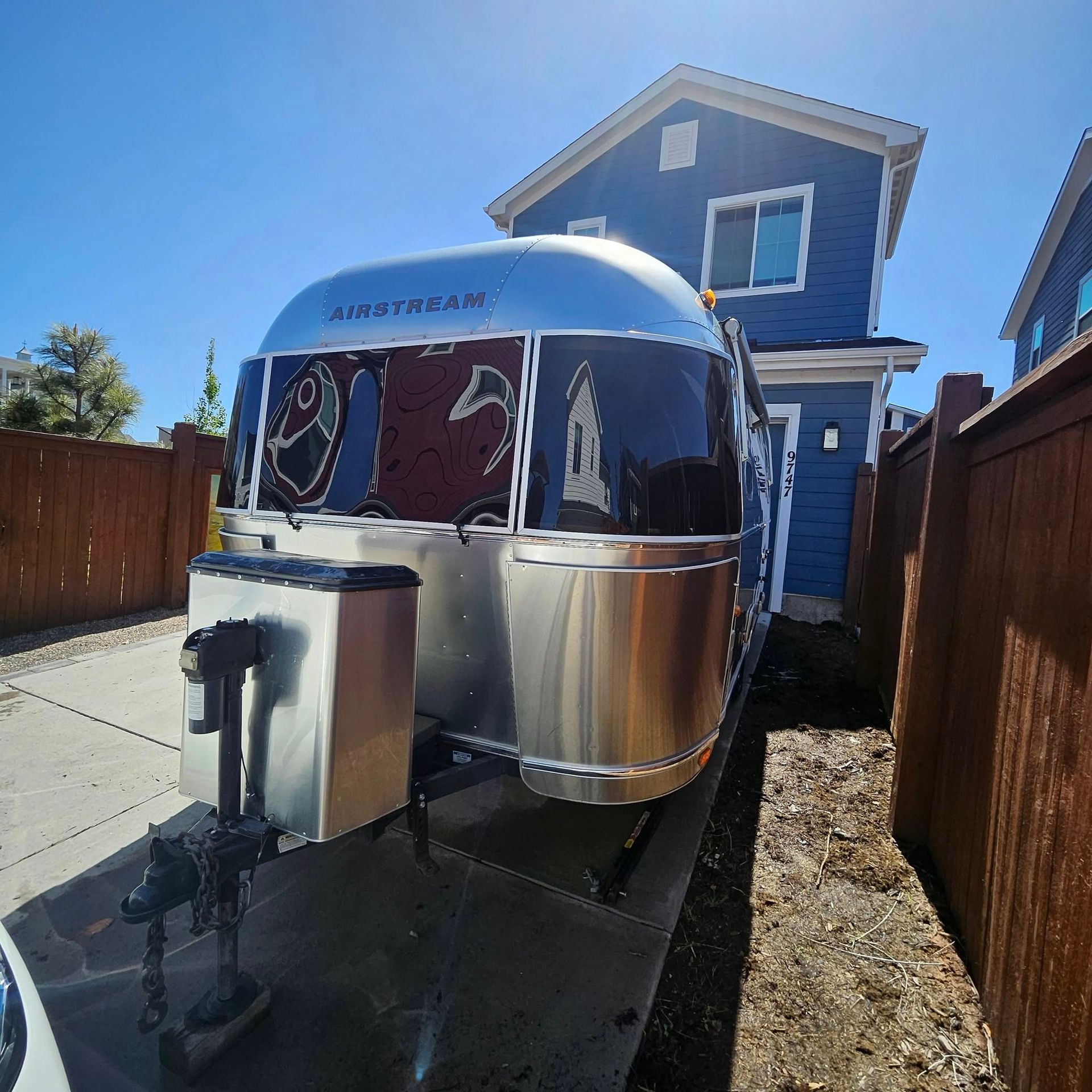 A silver Airstream travel trailer parked on a concrete driveway next to a blue house and a wooden fence.