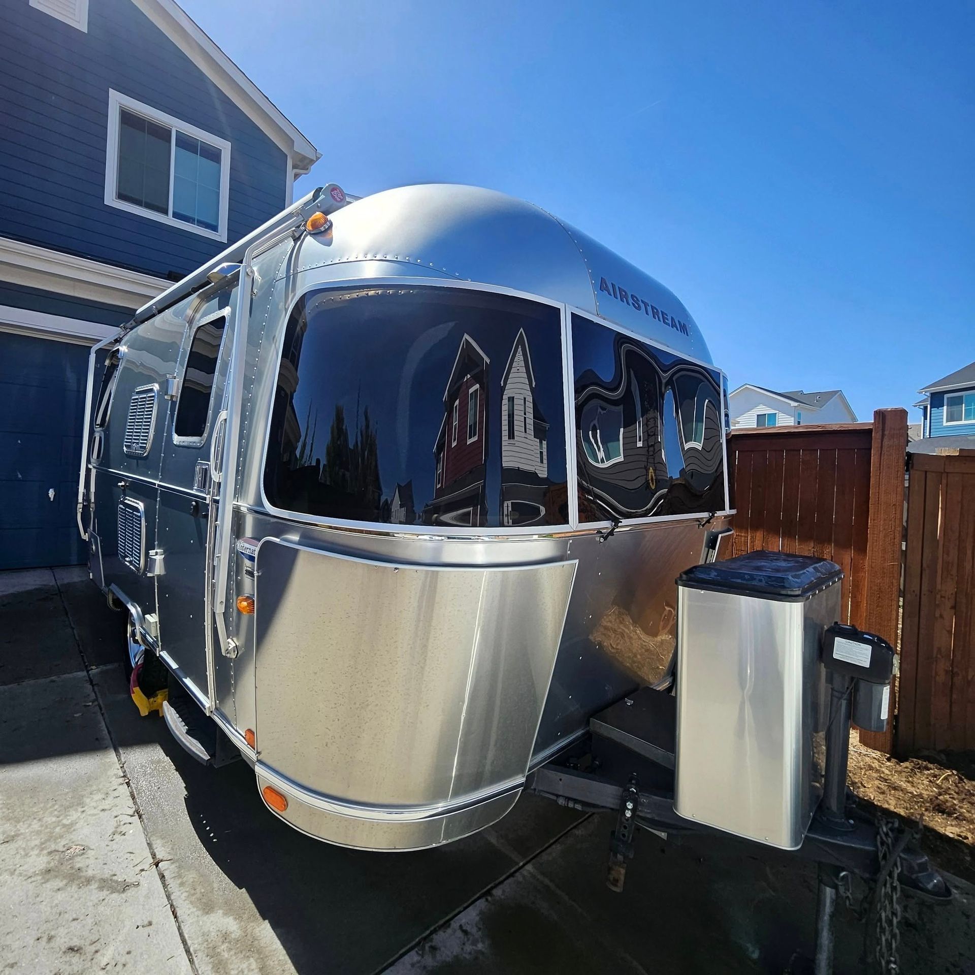 A polished silver Airstream travel trailer parked on a residential driveway next to a blue house and a wooden fence.