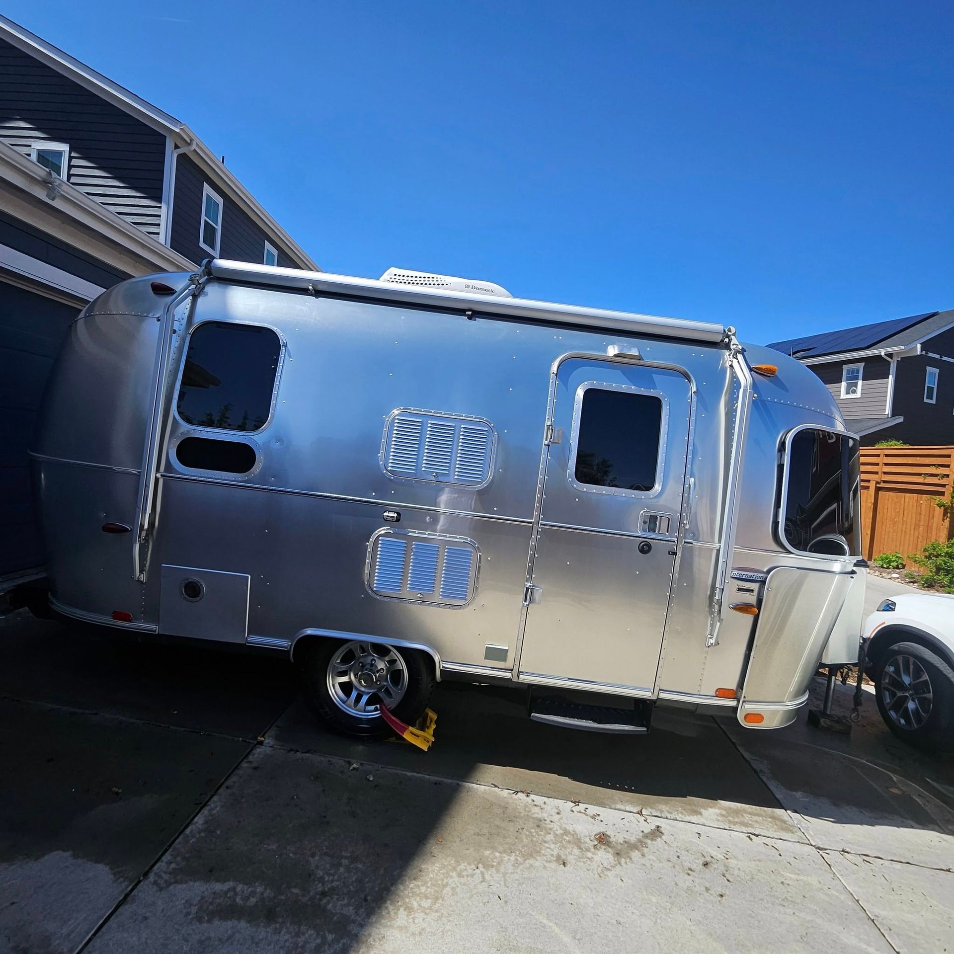 A silver Airstream travel trailer parked on a residential concrete driveway next to a house under a clear blue sky.