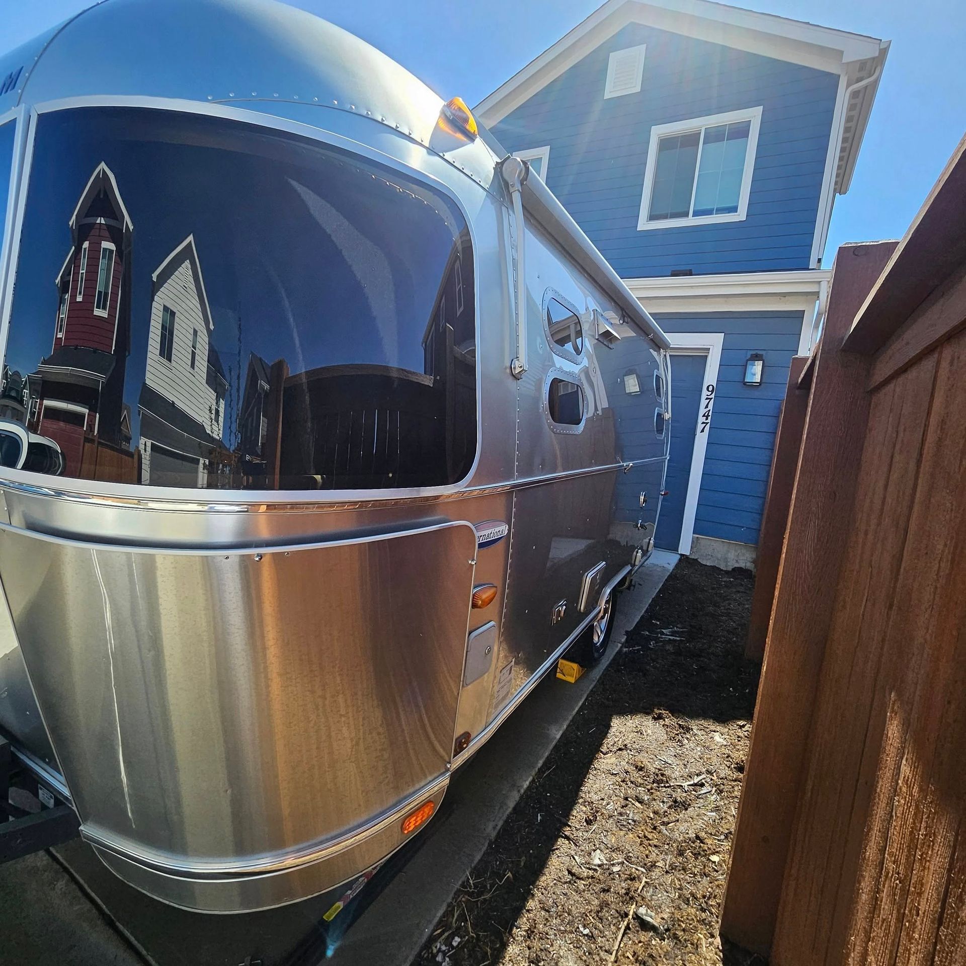 A shiny, rounded silver Airstream travel trailer parked on a paved driveway next to a blue house and a wooden fence.