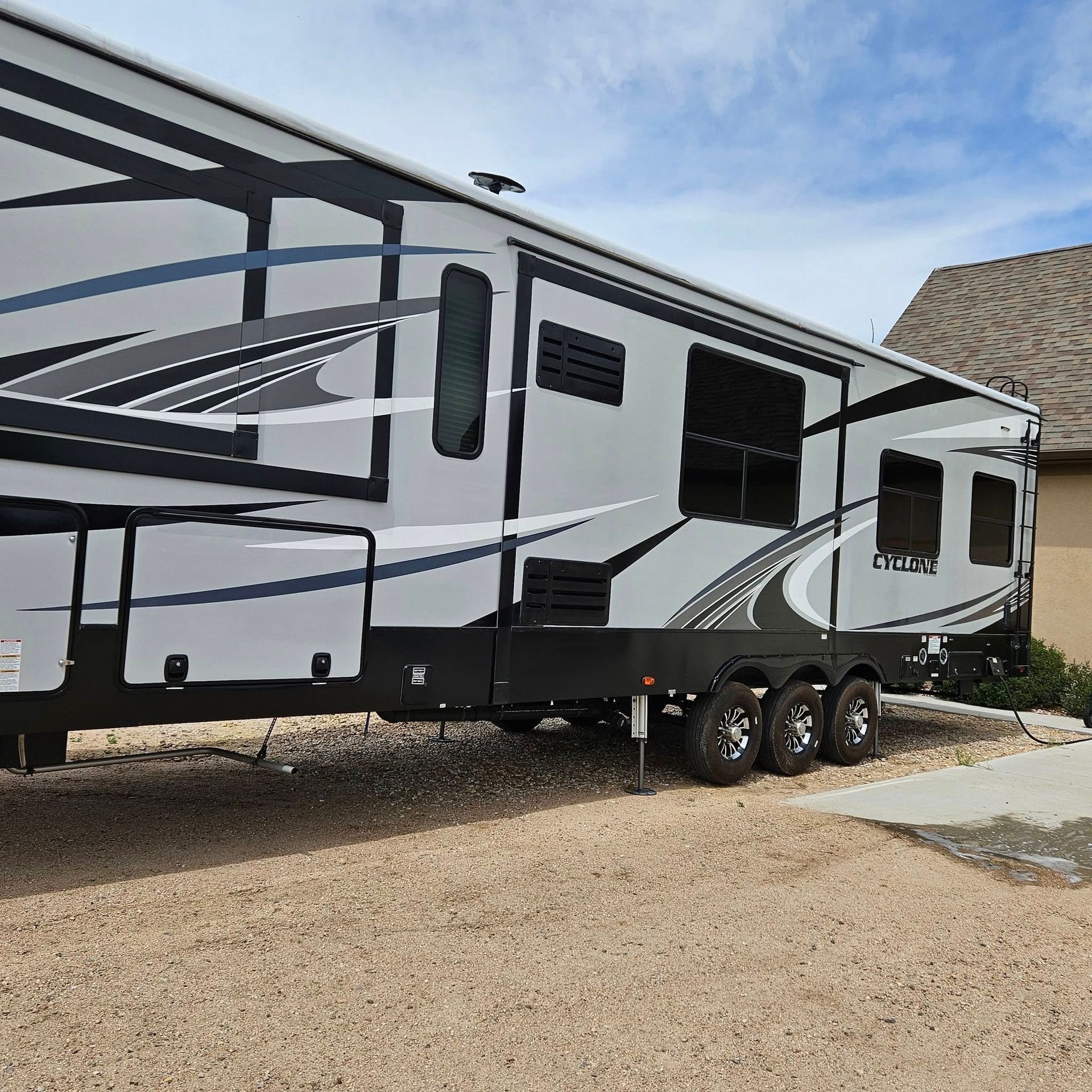 Side view of a parked RV with a gray and black geometric exterior design, sitting on a gravel driveway near a house.