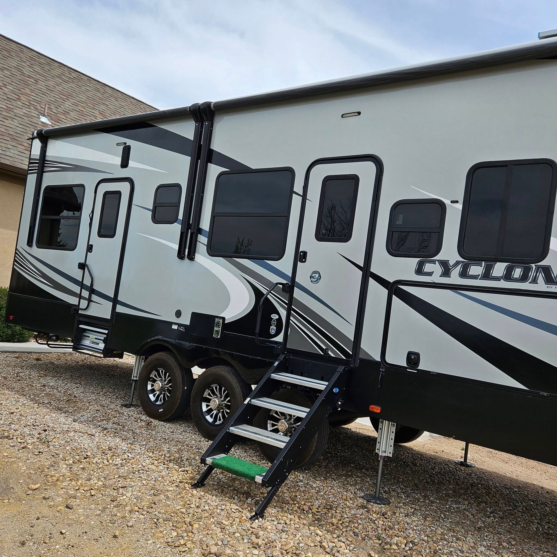A light gray and black Heartland Cyclone toy hauler RV parked on a gravel lot with its entry stairs deployed.