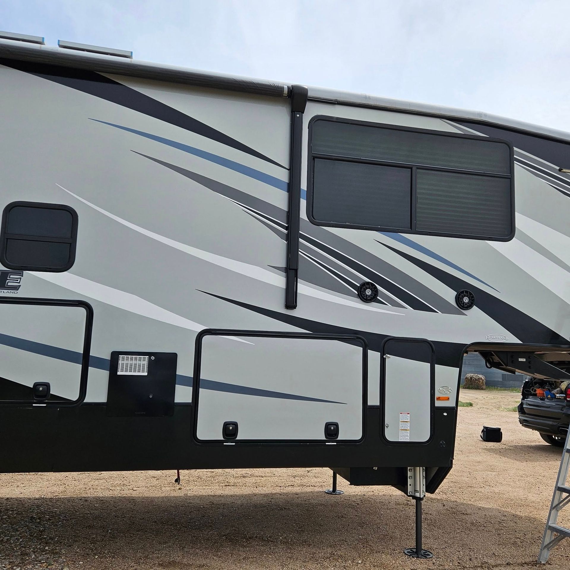 A side view of a fifth-wheel recreational vehicle with grey siding, dark blue graphic decals, and exterior storage doors.