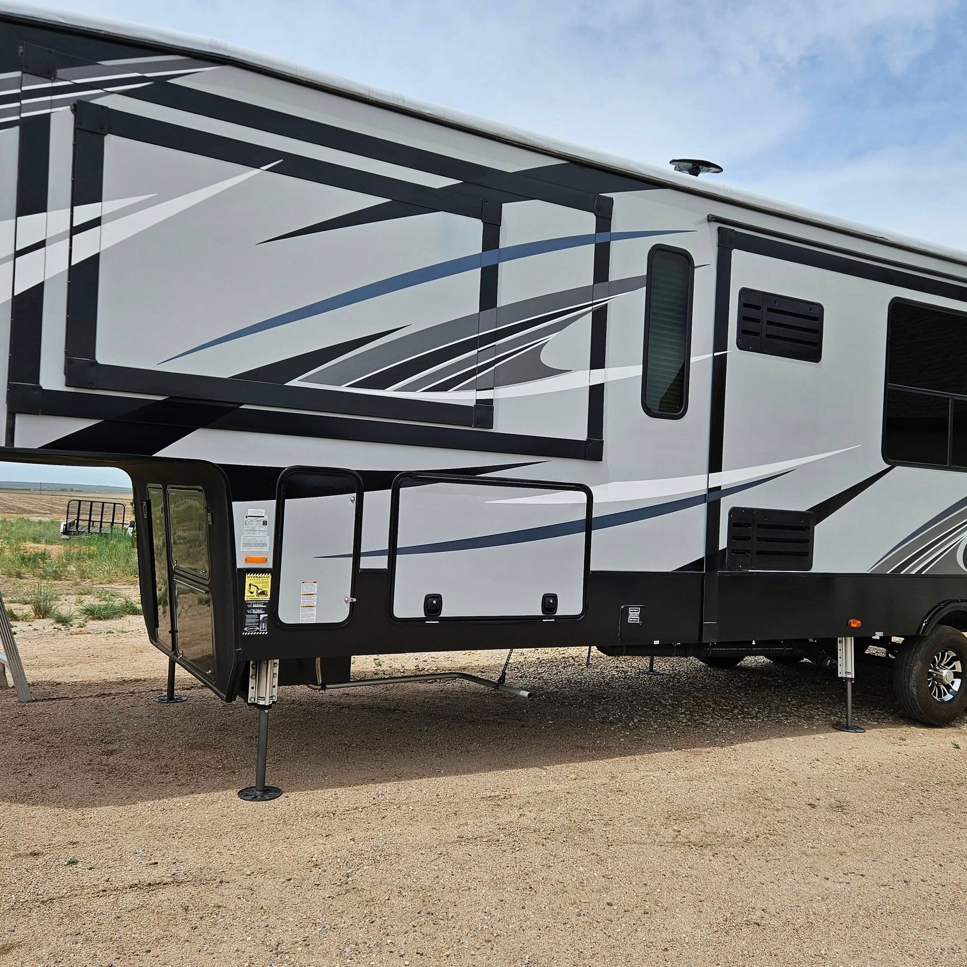 A fifth-wheel camper with a gray, white, and black geometric design parked on a gravel lot under a blue sky.