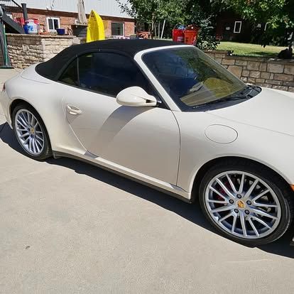 A cream-colored Porsche convertible with a black soft top parked on a concrete driveway in front of a house.