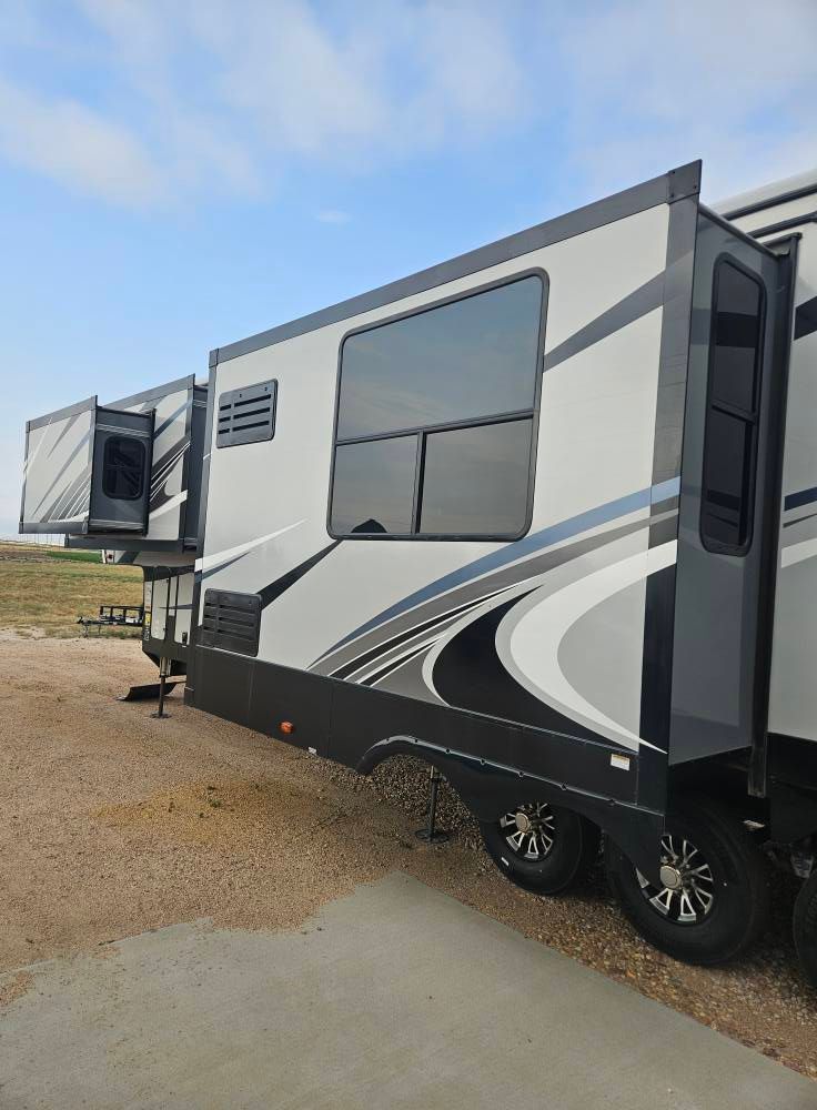 A parked RV featuring extended slide-outs, light gray walls with dark gray graphic accents, and black wheels on gravel.