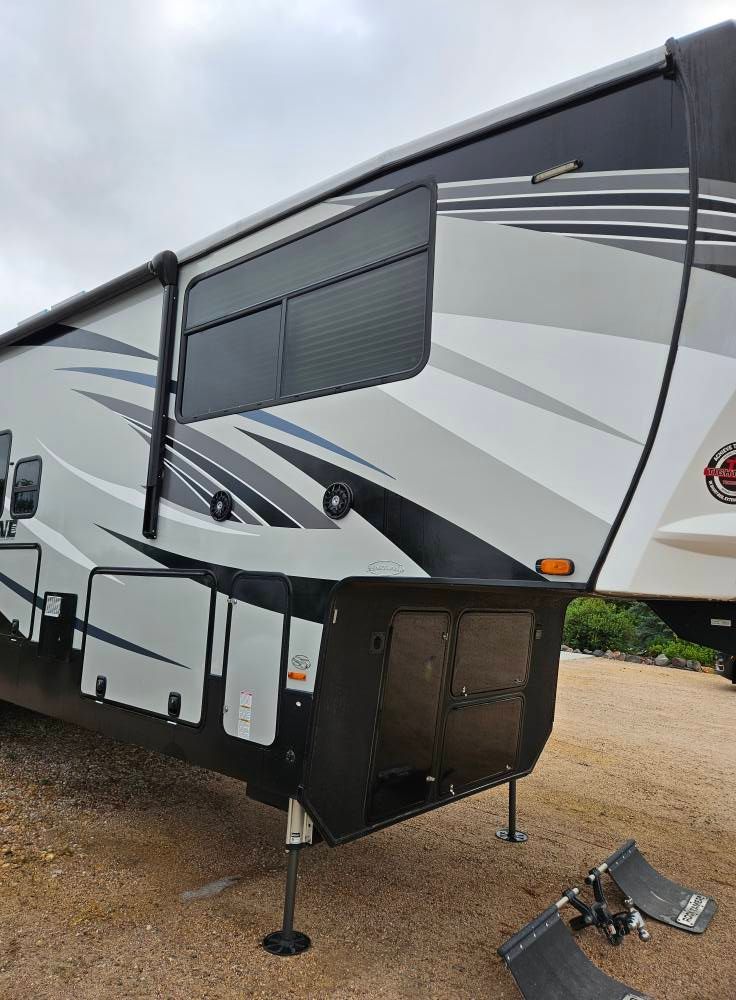 A white and gray fifth-wheel RV parked on a gravel lot with its landing gear deployed.