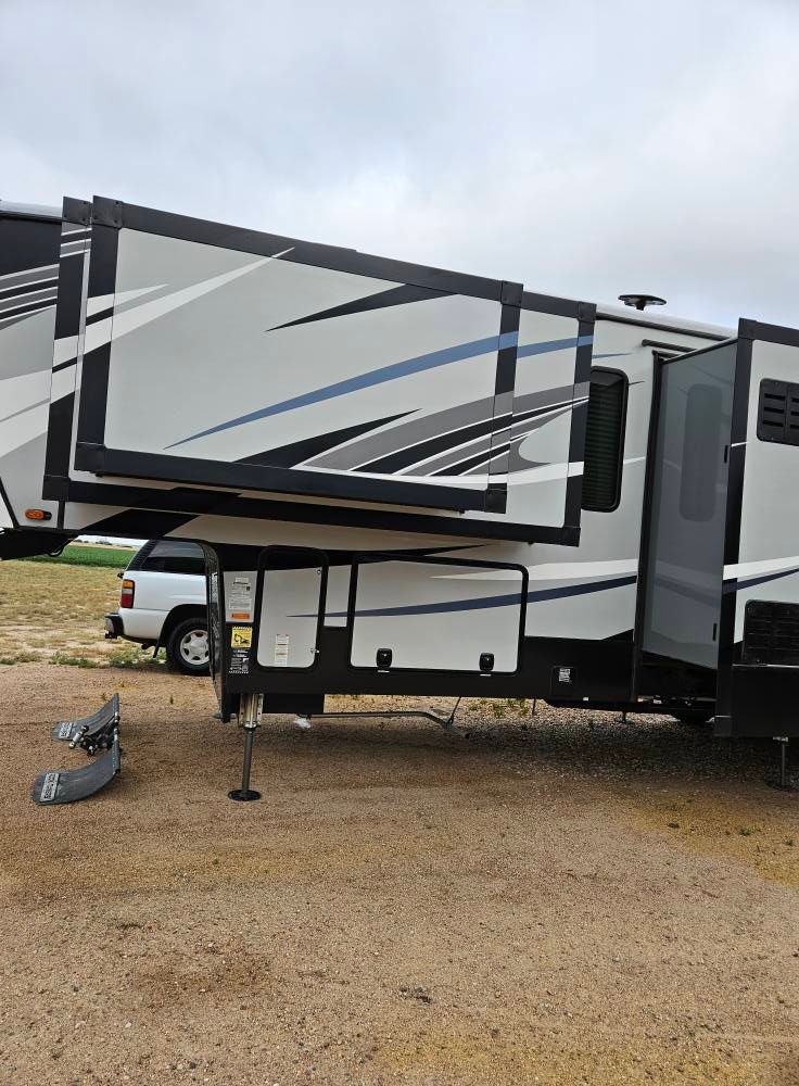 A side view of a large, modern fifth-wheel RV parked on a gravel lot, featuring an extended slide-out and white truck.