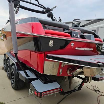 The rear view of a red and black motorboat on a multi-axle trailer, featuring a swim platform and overhead wakeboard tower.