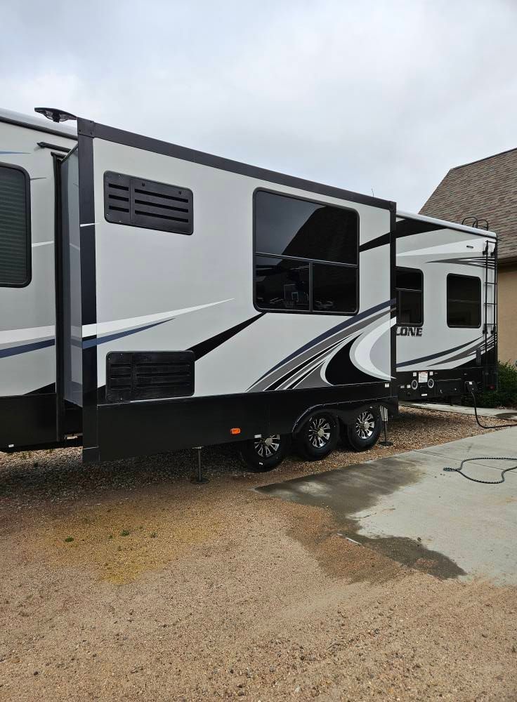 A white and black recreational vehicle with extended slide-outs parked on a gravel driveway next to a house.