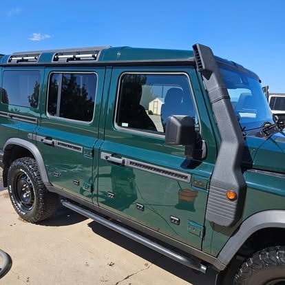 A dark green off-road vehicle with a snorkel intake on the passenger side, parked on a paved lot under a blue sky.