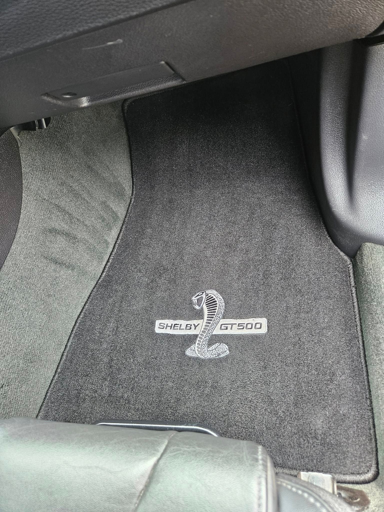 A dark gray car floor mat with a white embroidered Shelby Cobra snake logo positioned in the center.