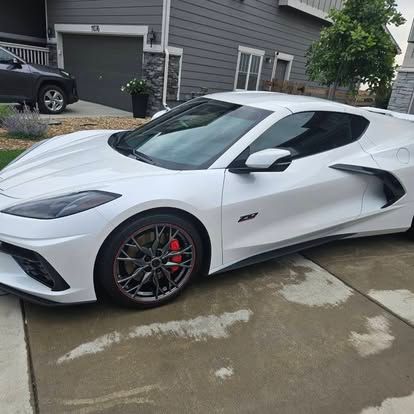 A white Chevrolet Corvette sports car parked on a suburban driveway in front of a gray house.