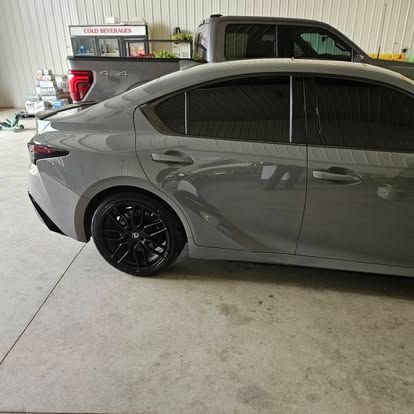 A side view of a gray sedan with black rims parked indoors, with a truck visible in the background.