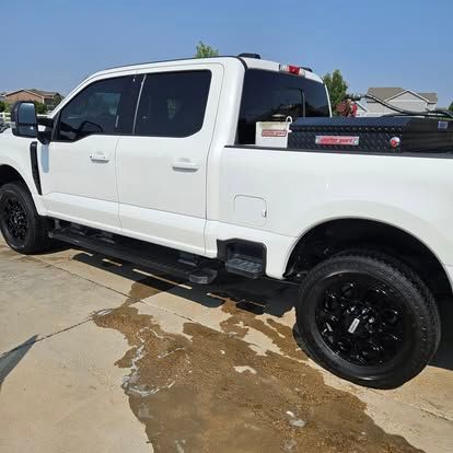 A white crew cab pickup truck with black wheels and a black storage box in the bed, parked on a concrete driveway.