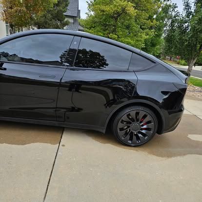 A black Tesla Model Y parked on a concrete driveway with tinted windows and black alloy wheels featuring red brake calipers.