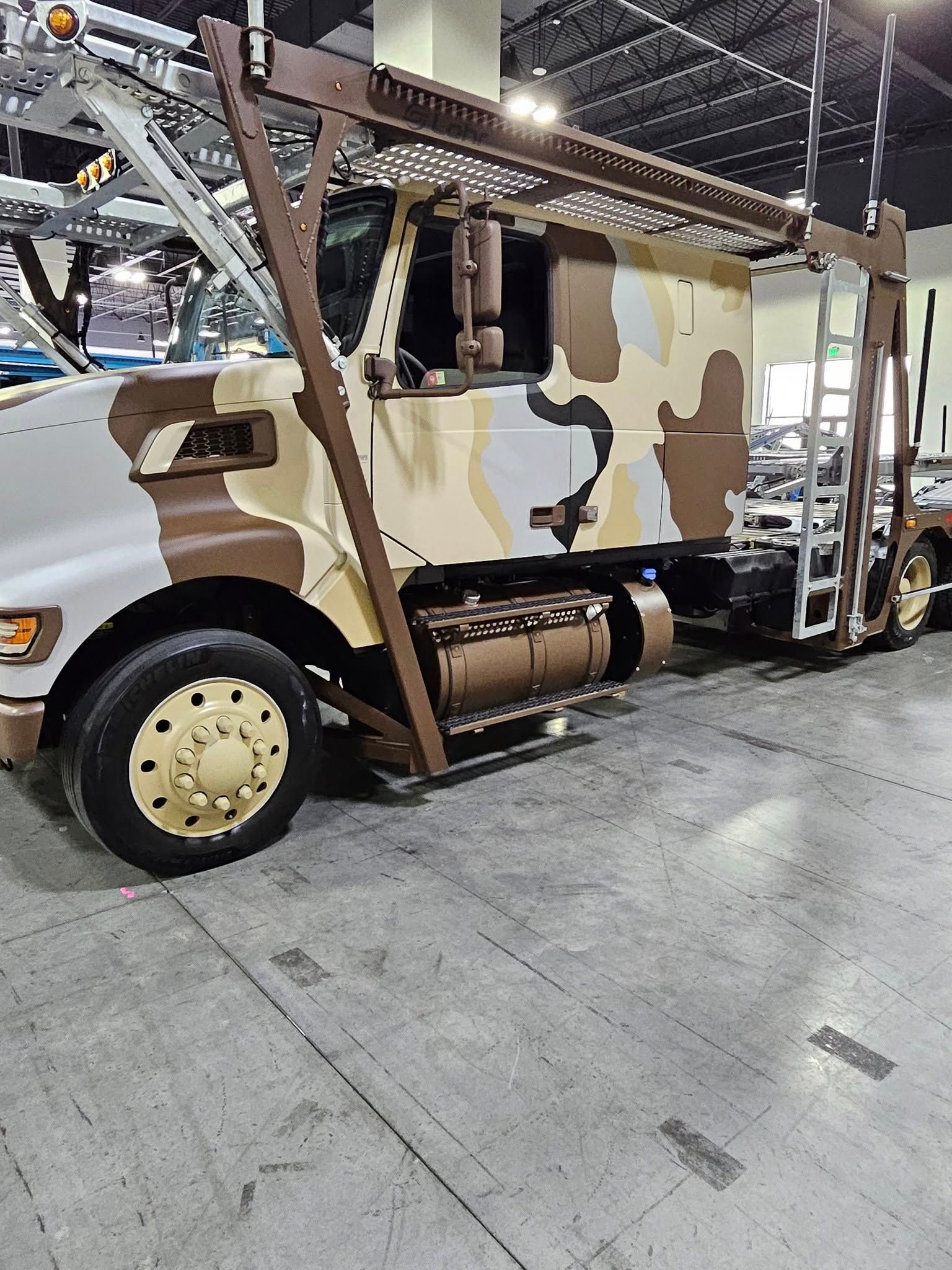 A semi-truck with a desert camouflage paint job and an attached metal rack frame, parked on a gray concrete floor.