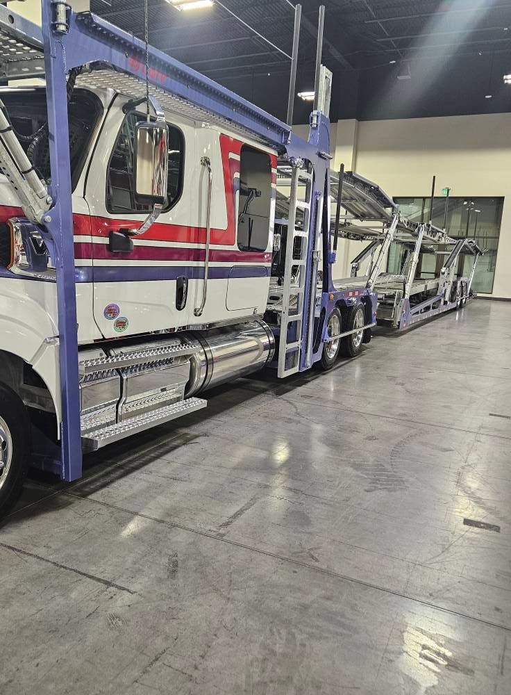 A white and blue semi-truck car hauler parked inside a spacious, brightly lit warehouse with a concrete floor.