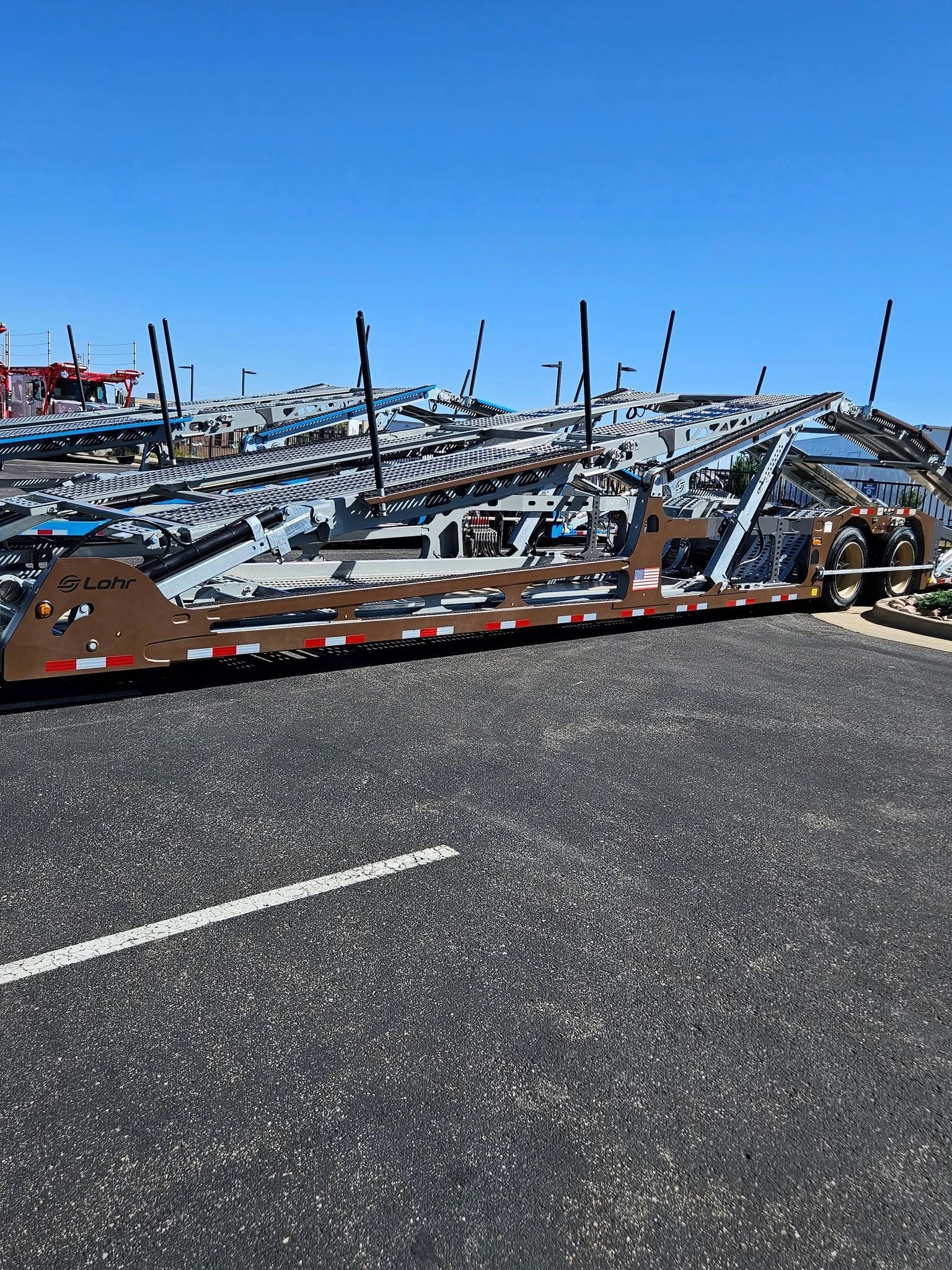 An empty brown metal car-hauler trailer parked on an asphalt lot under a clear blue sky.