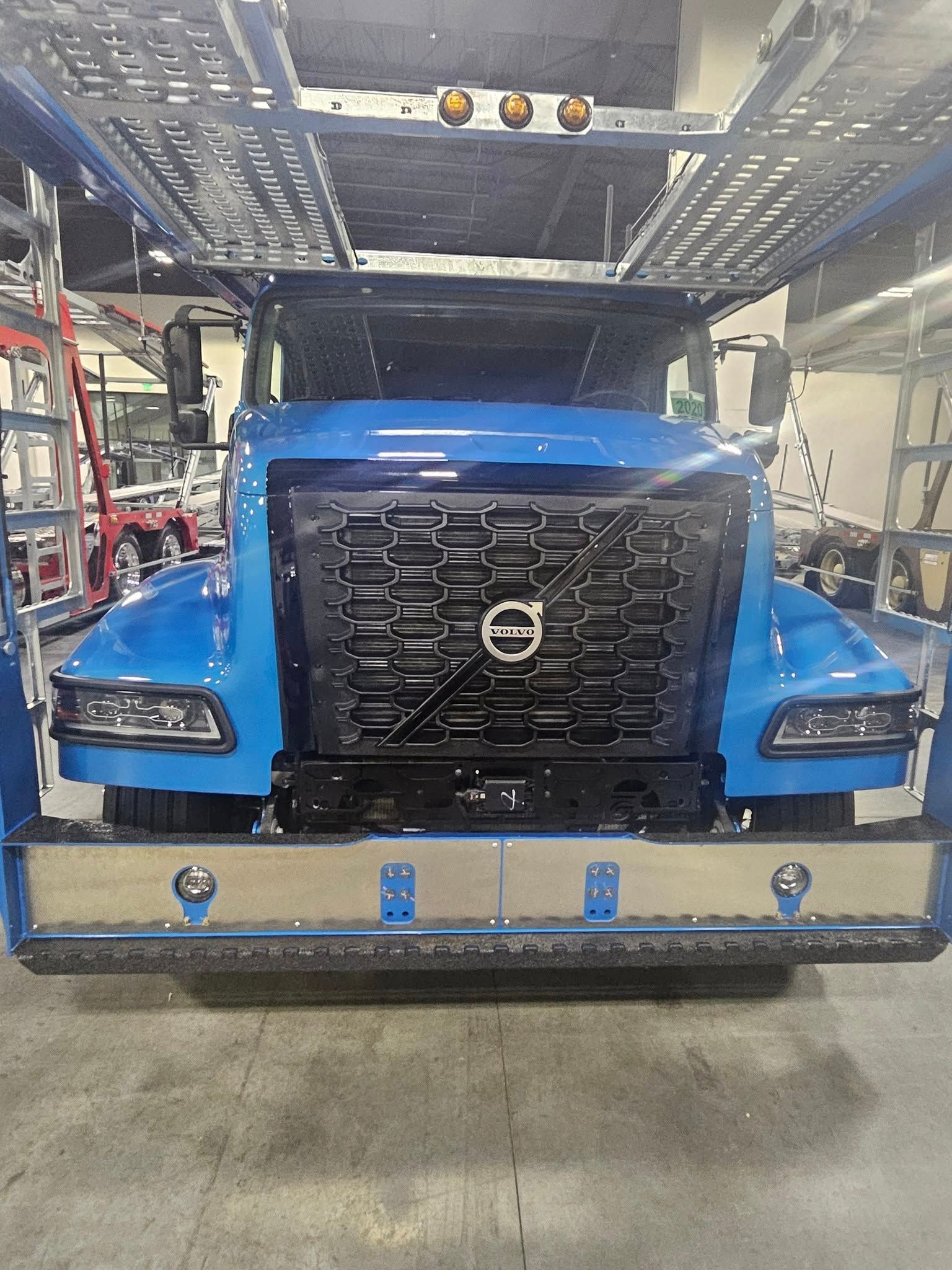 A front view of a bright blue Volvo semi-truck with an auto-carrier rack, parked inside a garage.