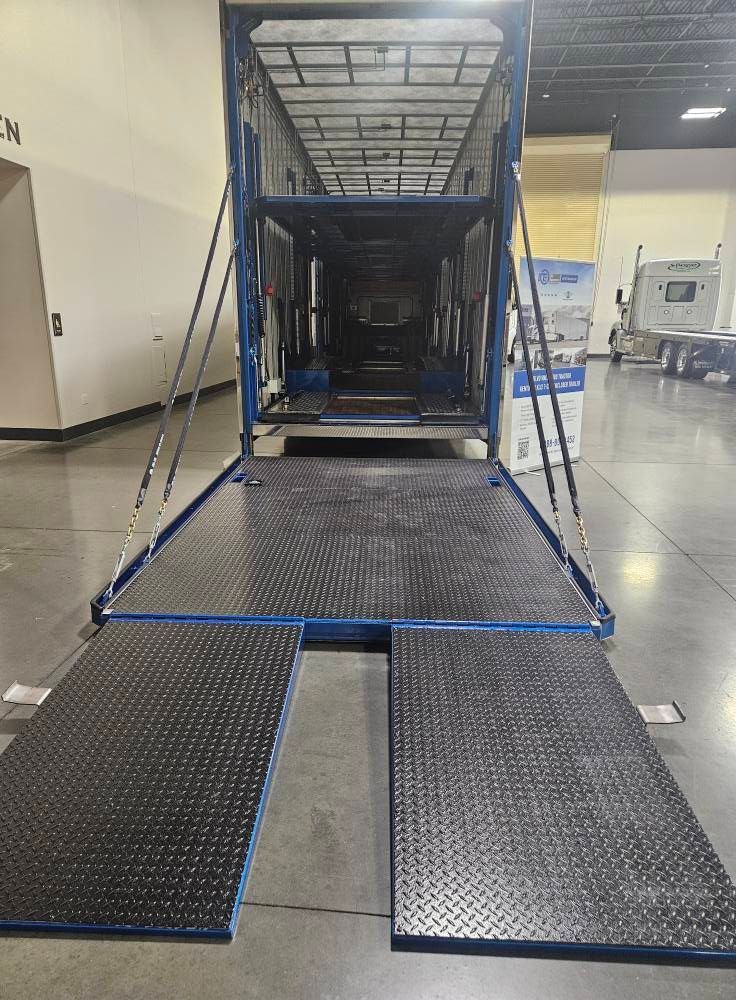 A blue car hauler trailer with a lowered, extended metal ramp at a loading bay inside a warehouse.
