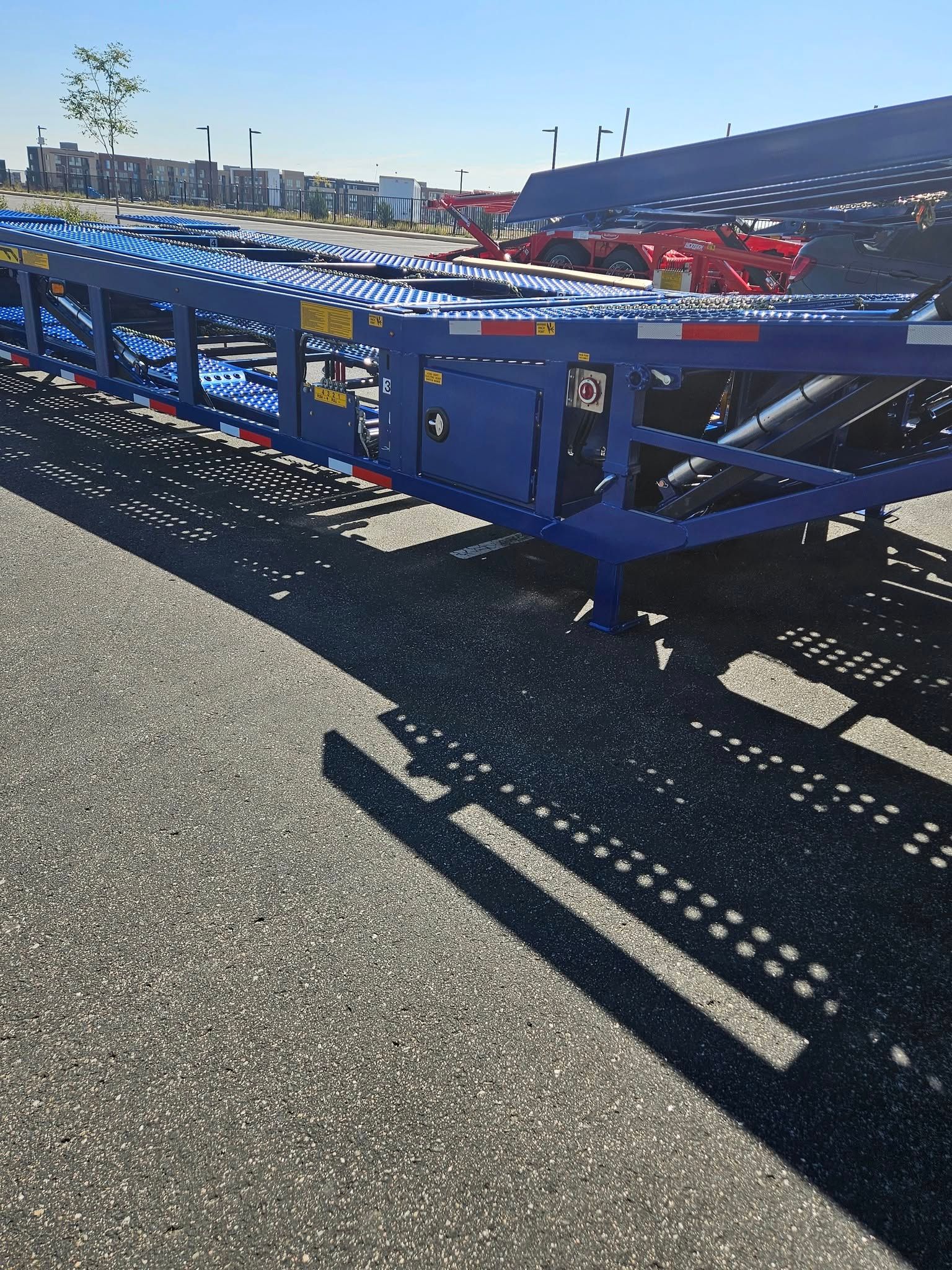 A blue metal car hauler trailer parked on an asphalt lot in bright sunlight, with a red truck visible in the background.