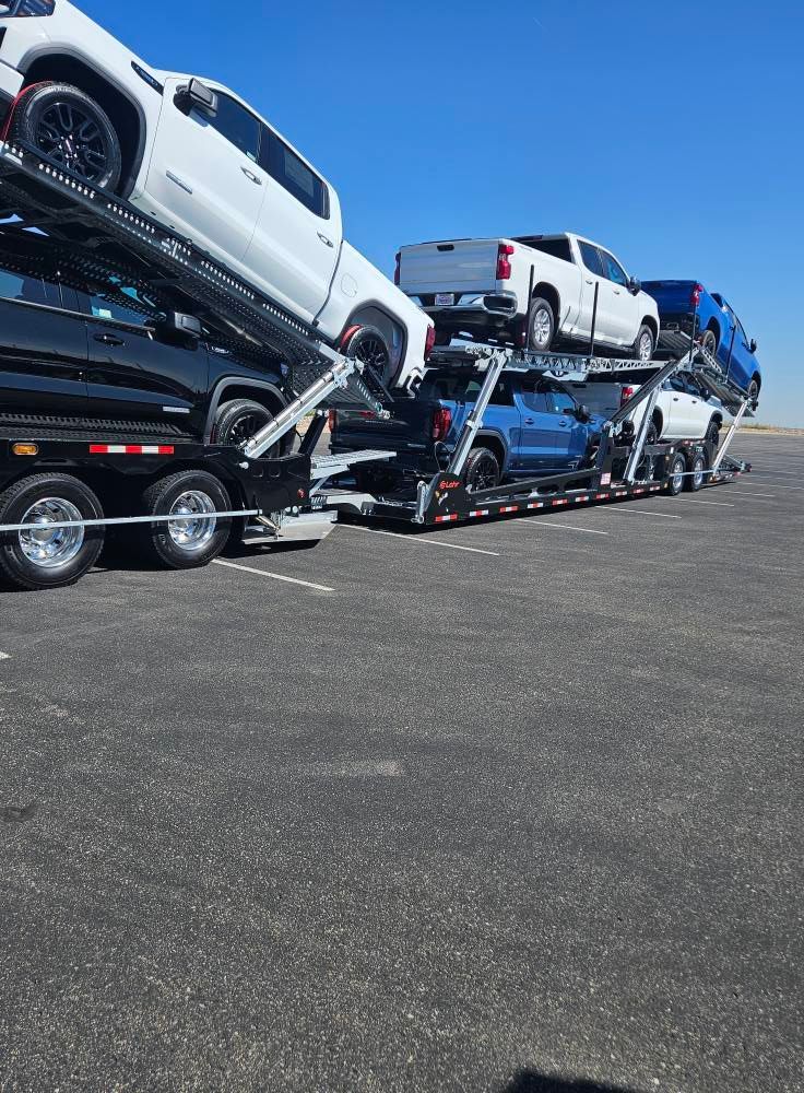 A car carrier trailer transporting multiple new pickup trucks of various colors across a flat, open lot under a blue sky.
