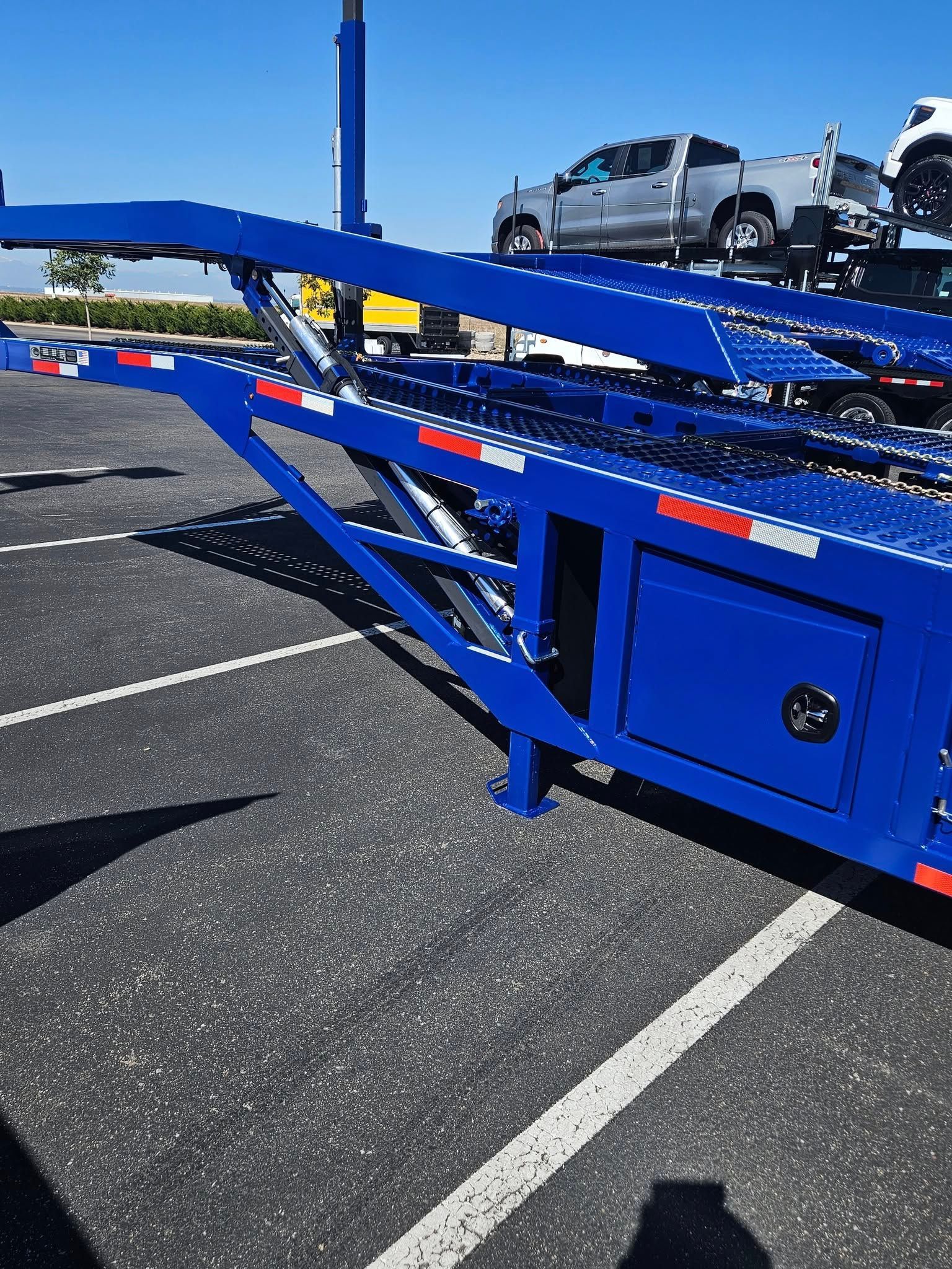 A bright blue car hauler trailer parked on asphalt, featuring a lowered ramp and a pickup truck loaded on the top deck.