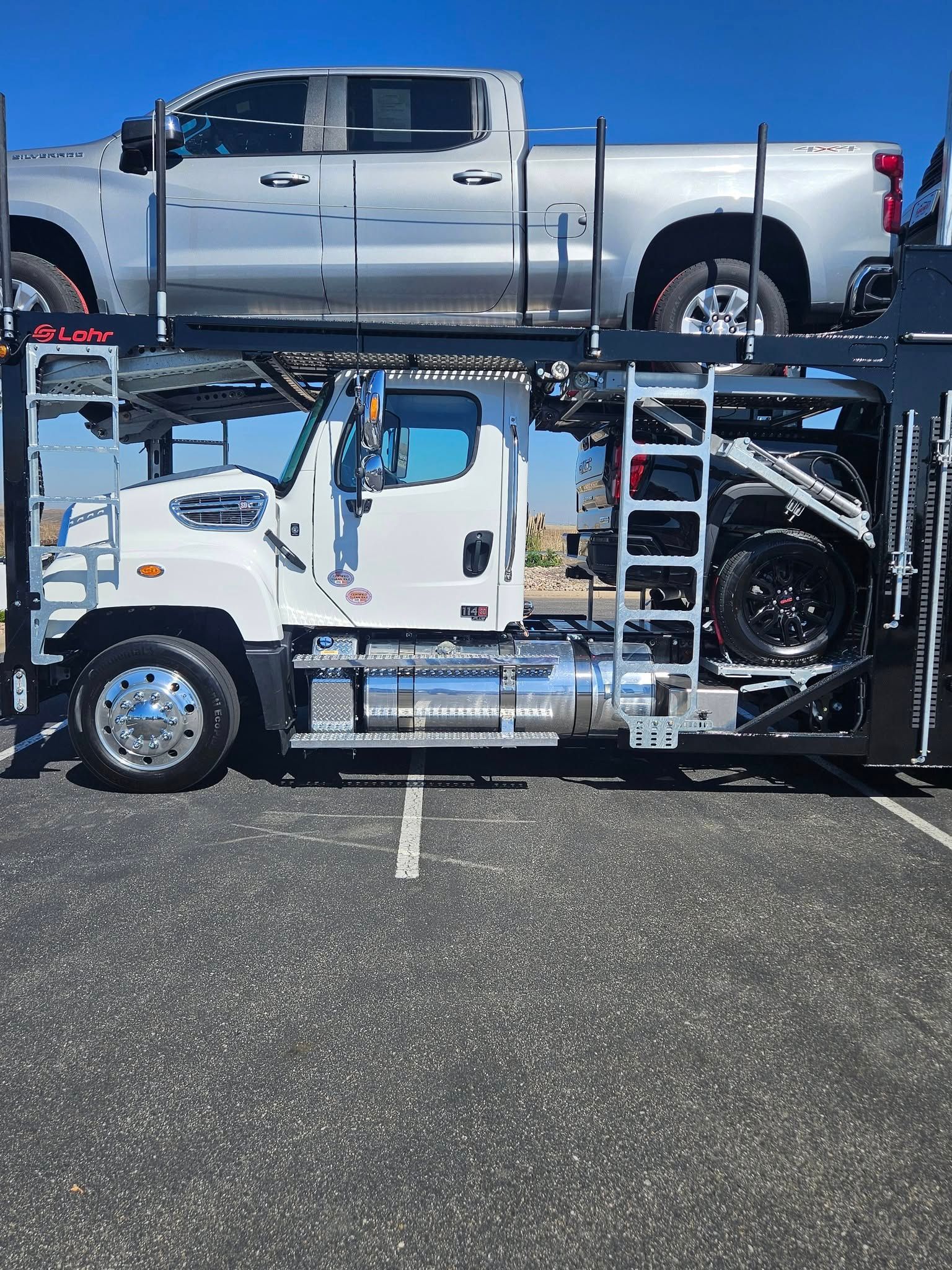 A car hauler truck parked on asphalt carries a silver pickup truck on its top deck and a dark vehicle on its lower level.
