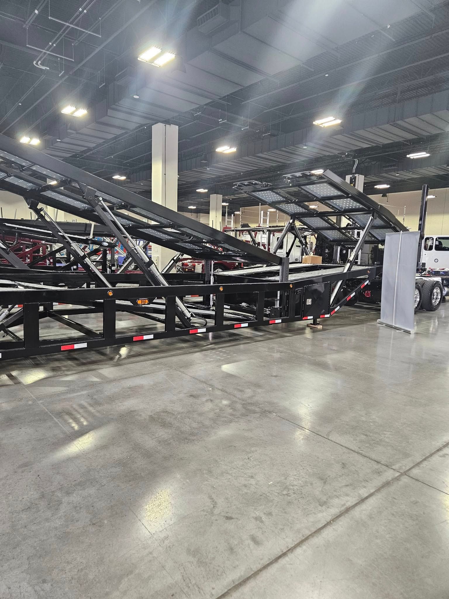 A black metal car hauler trailer standing on a polished concrete floor inside a large, illuminated indoor exhibit hall.