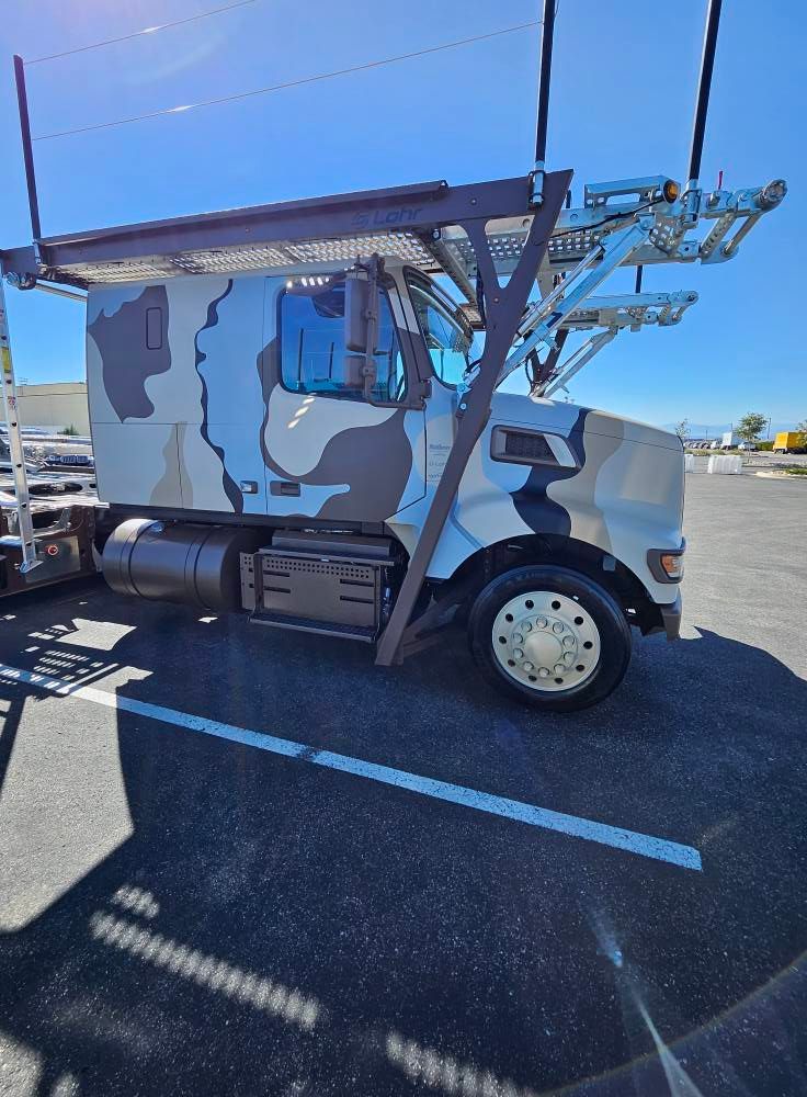 A camouflage-patterned car carrier truck parked in an asphalt lot under a clear blue sky.