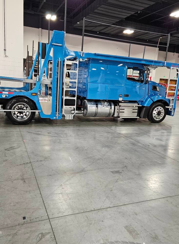 A bright blue semi-truck with an integrated car-hauler trailer parked inside a warehouse with polished concrete floors.