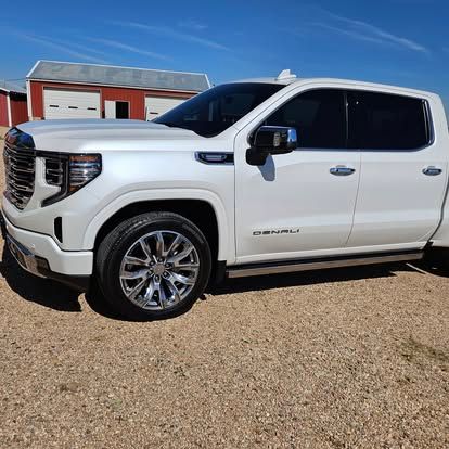 A white GMC Sierra Denali pickup truck parked on a gravel lot in front of a red barn under a clear blue sky.
