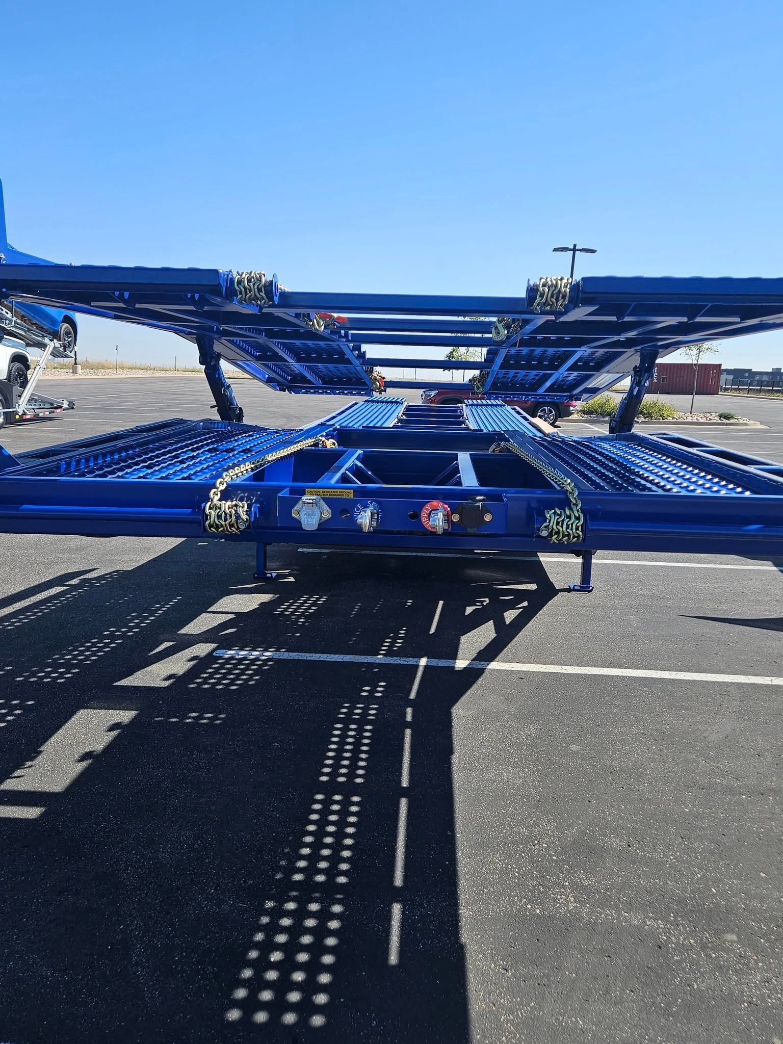 A blue, two-level car hauler trailer parked on asphalt in an open outdoor area under a clear blue sky.