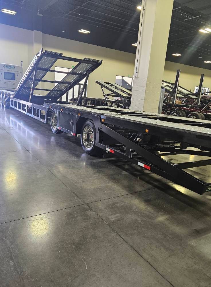 A black car hauler trailer parked inside a large, industrial warehouse space with concrete floors and bright lights.