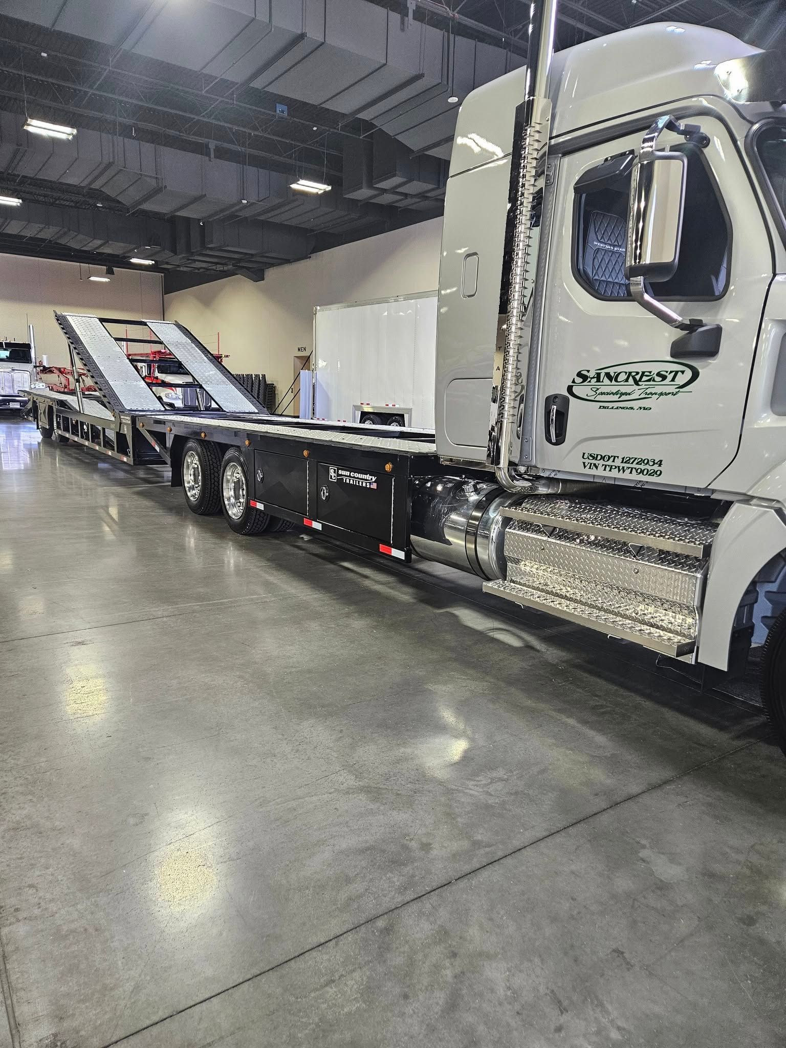 A white semi-truck with a black car-hauler trailer parked inside a large, brightly lit warehouse with a concrete floor.