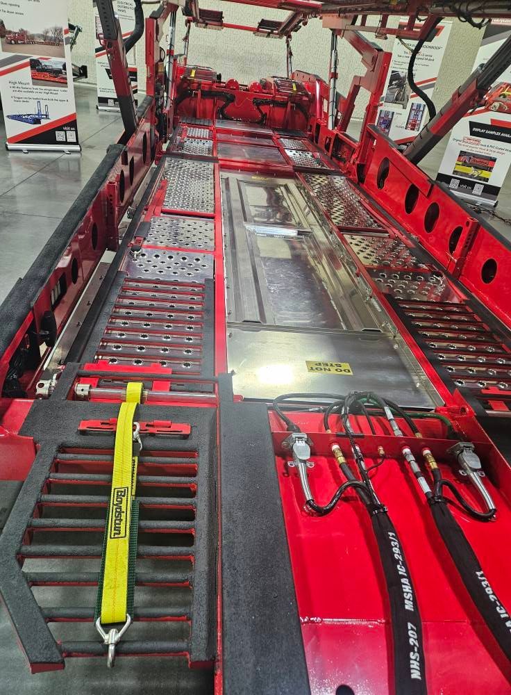 A high-angle view of a red car hauler trailer deck featuring metal grating, hydraulic lines, and a yellow tie-down strap.