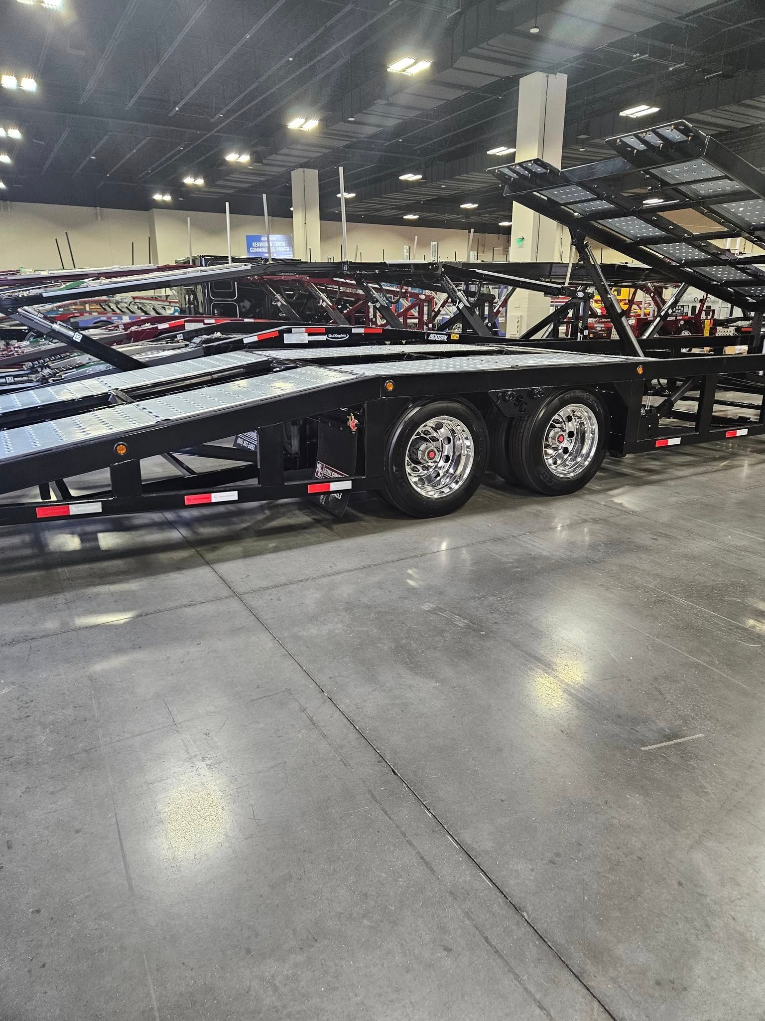 A black multi-level car hauler trailer parked on a concrete floor inside a large indoor exhibition hall.