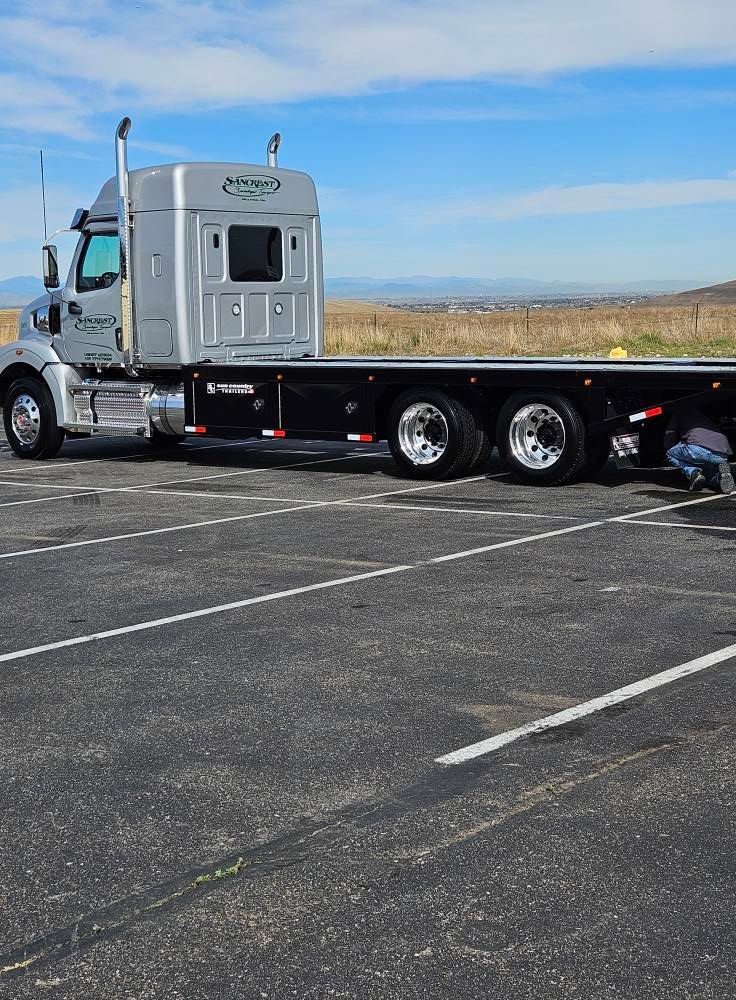 A silver semi-truck with a black flatbed parked in a large asphalt lot under a blue sky.