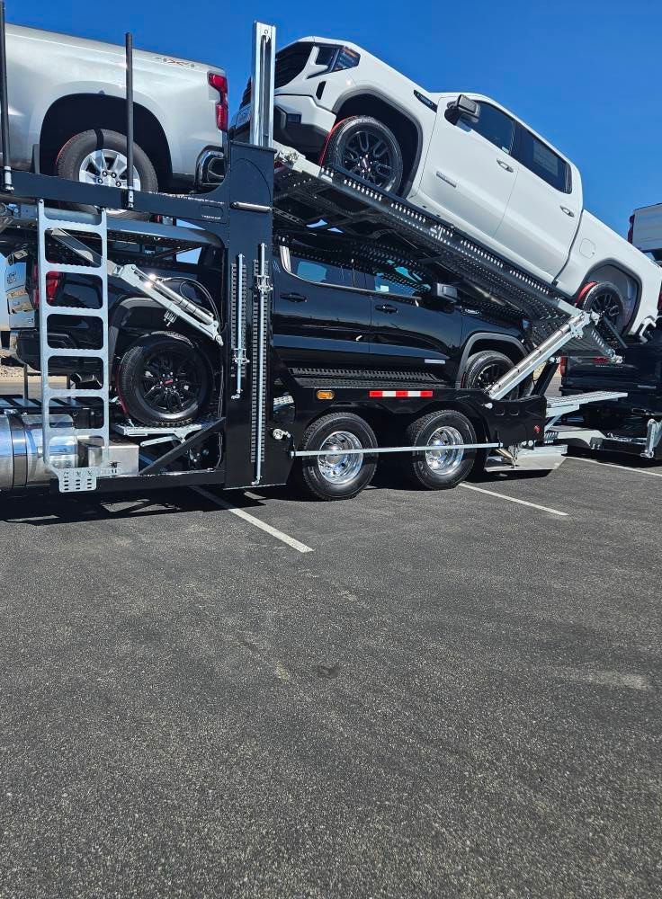 A car hauler trailer parked on asphalt, carrying three pickup trucks in white, black, and silver.