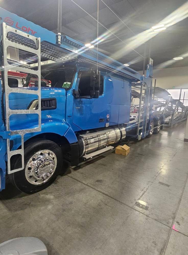 A bright blue semi-truck with a car-hauler trailer parked inside a large, illuminated indoor warehouse facility.