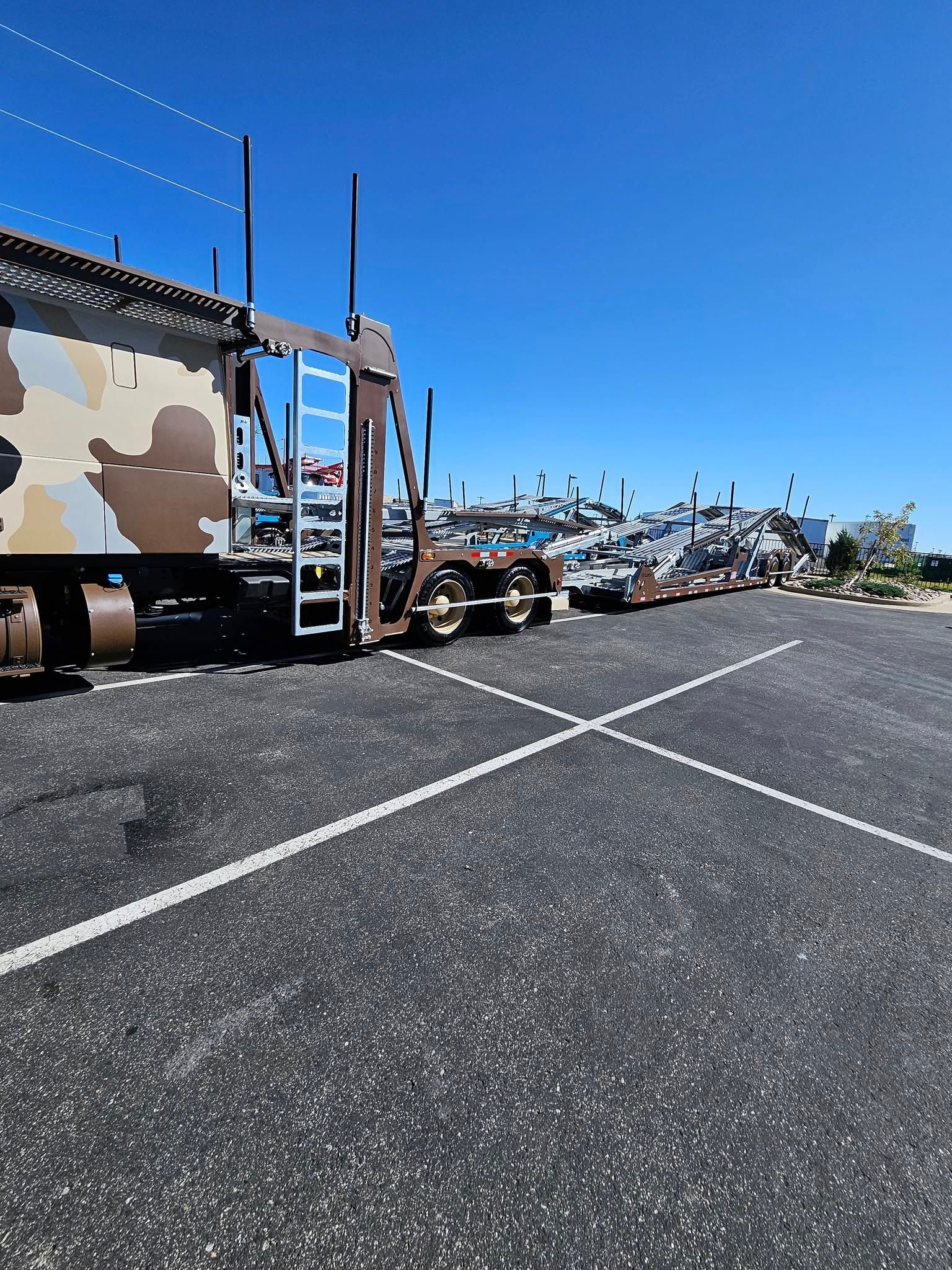 A camouflage-patterned car carrier trailer parked on an asphalt lot under a clear blue sky.