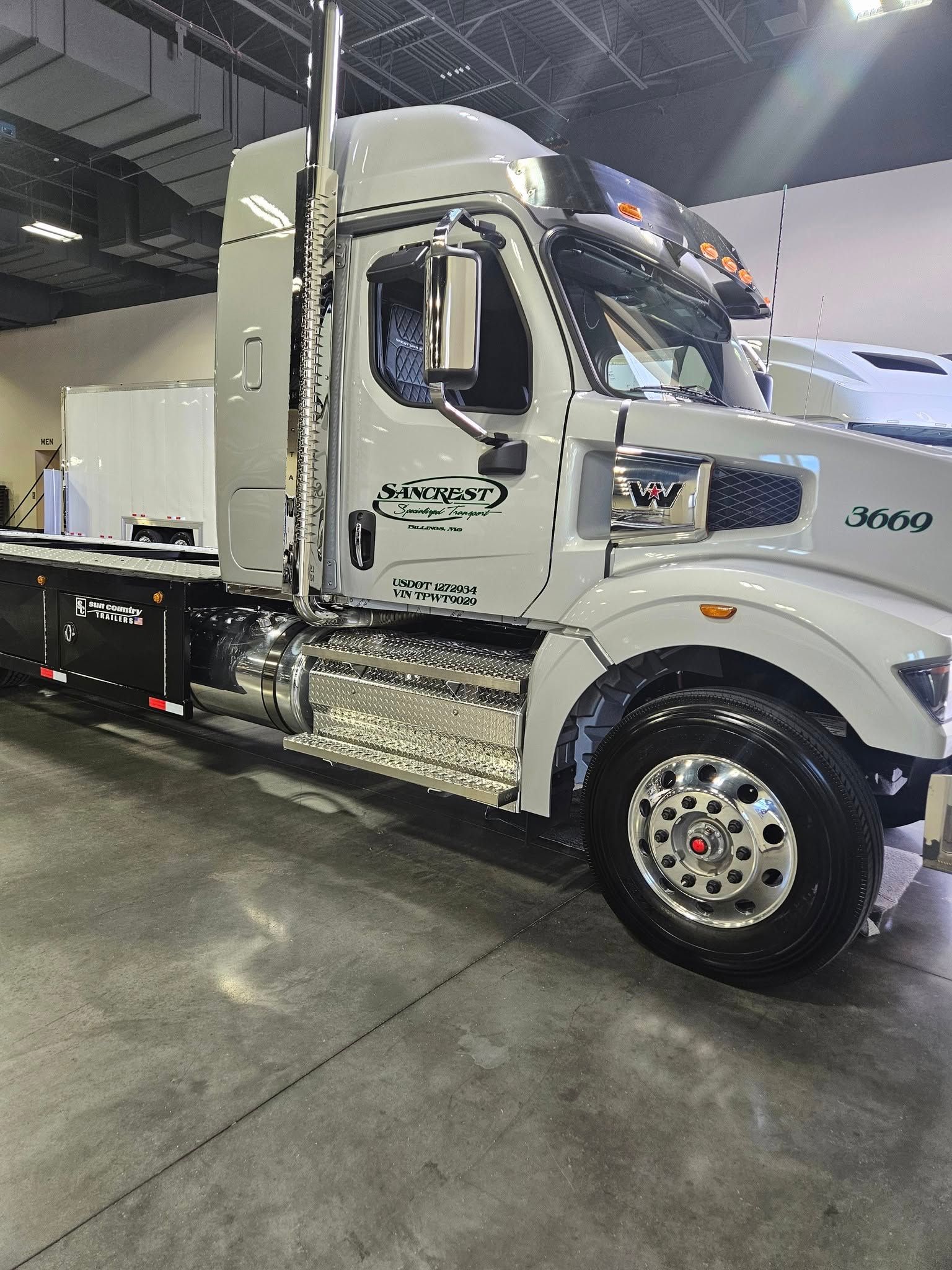 A light grey Western Star semi-truck parked indoors on a concrete floor, featuring chrome accents and side exhaust pipes.