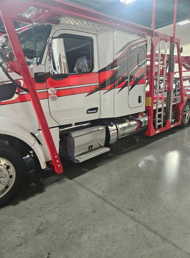 A white semi-truck with red and black graphic designs, hitched to an empty red car hauler trailer inside a garage.