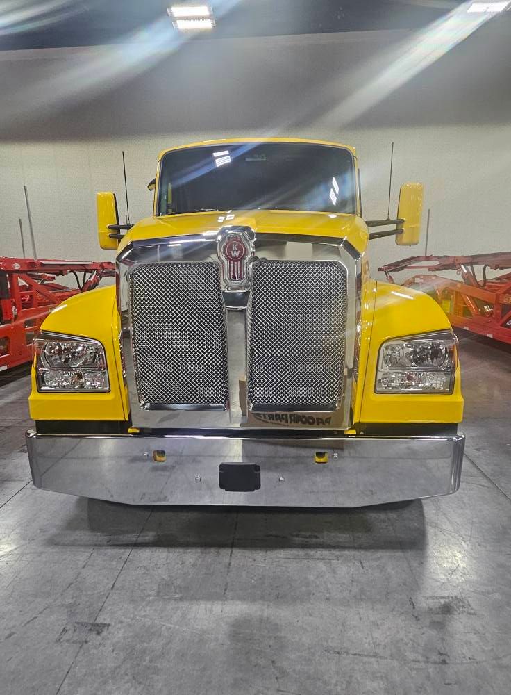 A bright yellow Kenworth semi-truck parked indoors, viewed from the front, featuring a prominent chrome grille and bumper.